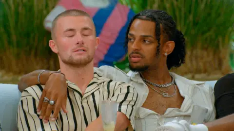A man sits with his eyes closed as another man drapes his arm around him.