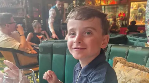 Photograph of Jacob, 8, pictured sat inside the Ivy restaurant. He sits on a green satin bench seat, has blue eyes and short brown hair and smiles at the camera. Behind him a table of customers can be seen, with a waiter leaned over their table reaching for a plate. 
