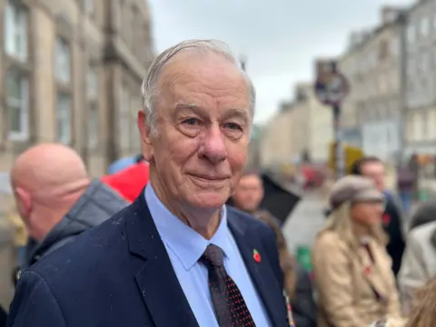 A older man with white hair wearing a blue blazer, a lighter blue shirt and a navy tie with red spots. He is wearing a red poppy brooch and stands in front of a crowd who are blurred in the background