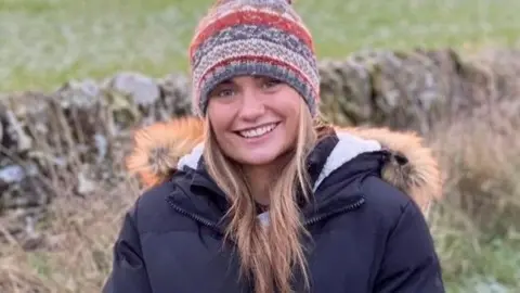 Hannah McInally, a young woman smiling at the camera, in a countryside setting, wearing a warm jacket and warm hat.