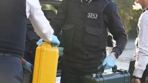 Serious Fraud Office officers wearing SFO-branded clothing and blue gloves carry hard cases, including a yellow case, beside an open car boot.