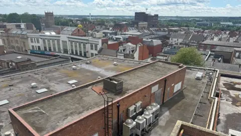 Annabel Amos/BBC Aerial shot showing roofs of commercial buildings with heating/air conditioning plant visible. The art deco white arcade building on Abington Street is visible to the left. The tower of St Giles Church is also visible to the left.