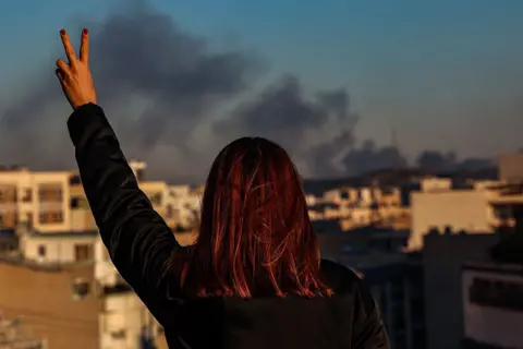 Getty Images A woman holds a two-finger peace sign, while she stands on a rooftop as smoke rises in the distance over Tehran on 3 March 2026.
