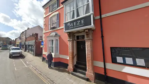 The front of a restaurant with pink walls, a brick porch and a black sign above the door which says Meze and a street with one car on it running alongside