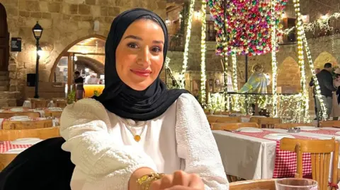 Supplied Alla Ghalayini is sitting in a restaurant covered in festive lighting. She is smiling and wearing a dark headscarf, with a white top and a gold necklace. She appears to be holding the hand of whoever is taking the picture. 