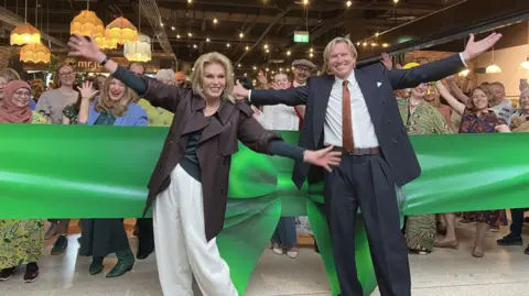 Joanna Lumley with a man in a dark suit both with their arms outspread in front of a huge green bow and lots of people behind in a shopping mall