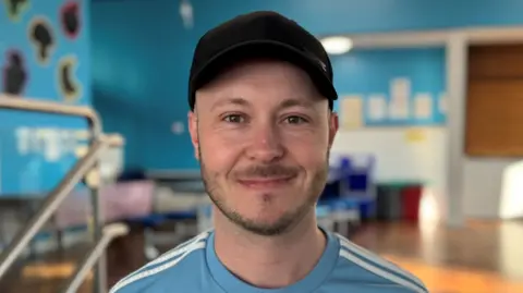 A man wearing a dark baseball cap and a light blue top with white stripes at the shoulders smiles into the camera. He has short dark stubble on his lips and chin. He stands in a room with blue walls and posters on the walls.