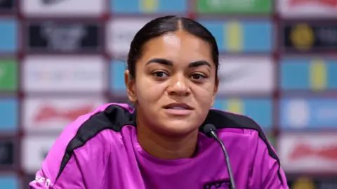 PA Media Footballer Jess Carter sits at a desk speaking into a microphone. She is wearing a purple and black jersey. 