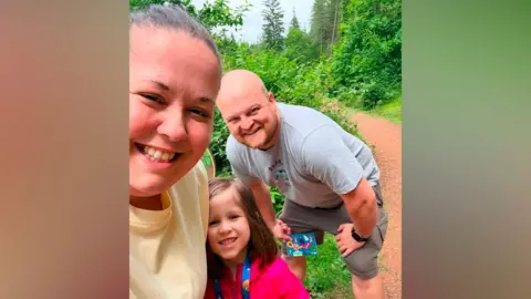 Handout A family of three - a woman, a man and their daughter - smile as they stand on a bark path through a lush green forest. The man, who is bald, is wearing grey shorts and a grey T-shirt and is crouching with his hands resting on his knees while his daughter, who has shoulder-length brown hair, is wearing a pink coat. His wife has slicked-back brown hair and is wearing a yellow T-shirt.