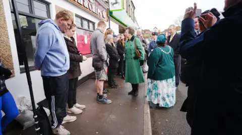 PA Media Princess Anne wearing a long green coat stands outside of a fish and chip shop. There is a row of people in front of her, and one person taking a picture.