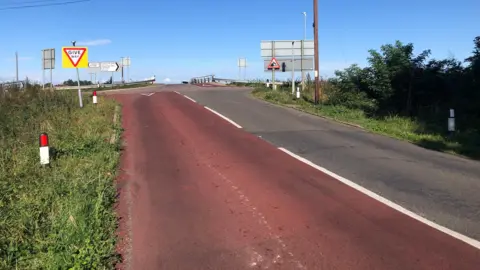 JOHN DEVINE/BBC Boots Bridge approach in Manea, Cambridgeshire, looking up an incline on red-painted tarmac towards the bridge, with a "give way" warning sign 
 and another "quayside or river bank" sign showing a vehicle plunging in to water