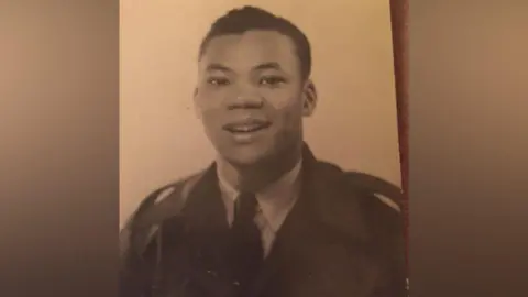 A black and white photo of a young man with short brown hair smiling for the camera and wearing a dark blazer, tie and light shirt. He looks very smart and is in uniform.