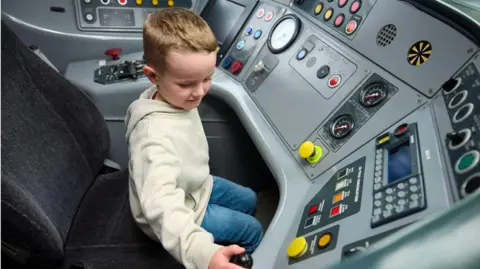 AvantiWestCoast Five-year-old Daniel Rogerson from Wigan sits in the driver's cab of one of his favourite trains. He is operating some of the buttons while sitting in the driver's seat. Daniel wears a cream jumper and blue denim jeans. 