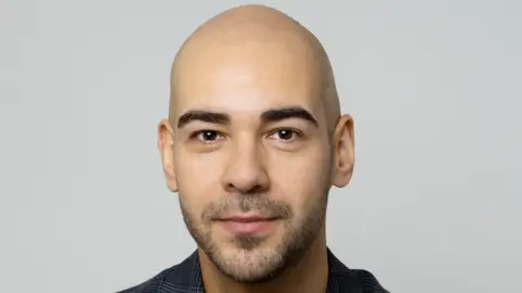 AeroGrid A bald man with stubble smiles without teeth at the camera wearing a black t-shirt and grey and white blazer. 