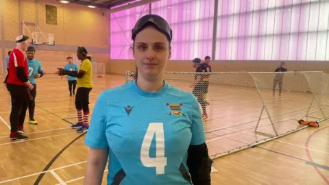 Goalball player Nansi Hidrej is wearing a blue sports top with a white number four on the front. She is stood in a sports hall with other players in the background. There are a set of goals and a wall which is illuminated with purple light.