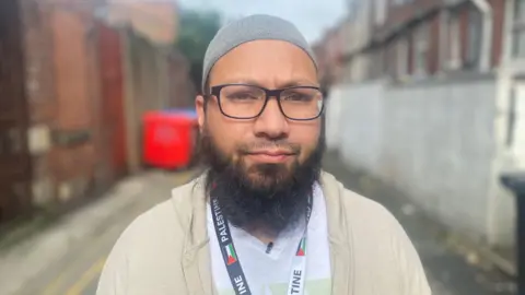 Mothin Ali, looking straight at the camera, and wearing black glasses and a kufi hat. 