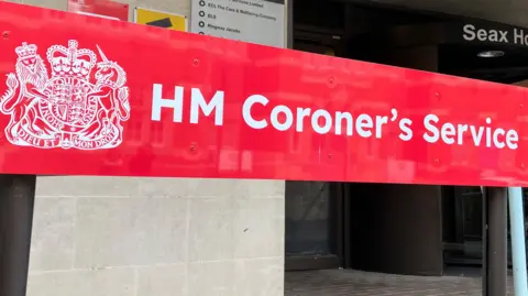 Lewis Adams/BBC A red sign that says HM Coroner's Service. It is at the entrance to Essex Coroner's Court, which is a cream building that has a black revolving door and brick paving outside.