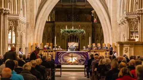 Les Pickersgill People attending a carol service inside Wells Cathedral. The pews are all filled with people and the choir is singing by the altar.