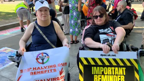 Two women using wheelchairs are in a public square. They are holding banners in front of them. The one on the left reads: 'Stop halving out benefits'. 