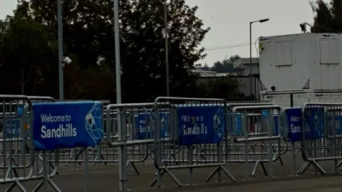 BBC News/Andy Gill A large number of metal barriers, some with "Welcome to Sandhills" written on them on blue banners. 