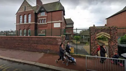 Google A general view of Sacred Heart Catholic Primary School in Barrow-in-Furness. It is an old red brick building with a brick wall topped with a metal fence around the playground. People are pushing buggies in front of the school.