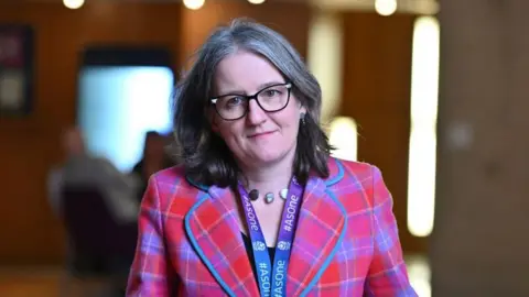 Maree Todd wearing a red and purple tartan jacket and large-rimmed glasses walking through the Scottish Parliament building