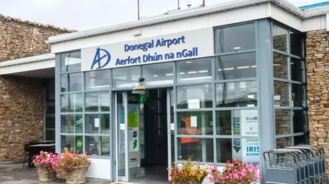 Getty Images The front of Donegal airport, it is a small building with a glass entrance porch and stone walls on either side. The sign on the porch reads "Donegal Airport" in blue font on a grey background, with the Irish translation underneath. 
