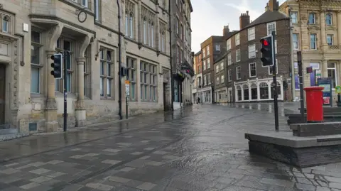 Claire Donlan Water flowing down an empty Market Square in Durham city centre. There are traffic lights on red and a red post box. 