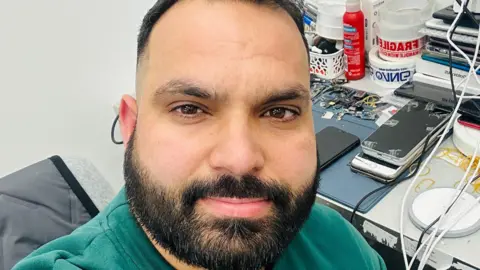Naveed Aslam, who has short, dark hair and a dark beard and wears a green sweater. He is sitting at a desk which has several mobile phones on it.