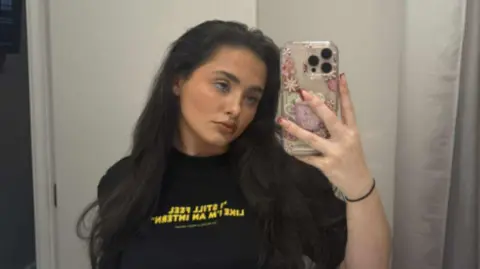 Charley Harrison Charley Harrison, with long brown hair and wearing a black t-shirt with yellow writing, holds her mobile phone in front of a mirror to take a selfie.