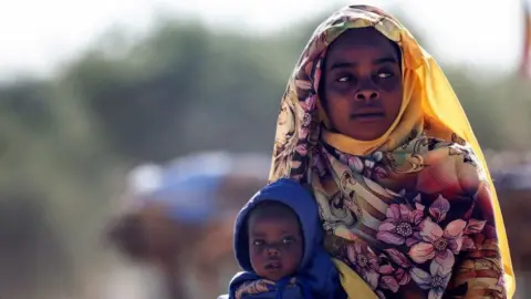 A woman in a yellow head scarf is seated behind a child in a blue hoody.