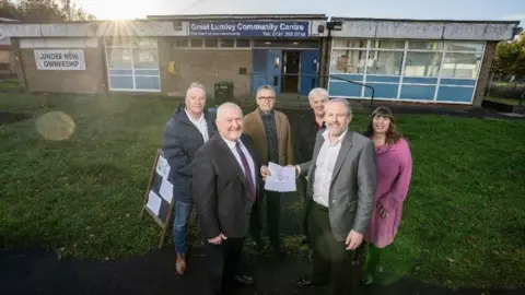 Phil Heaviside (front left) and Councillor Mark Wilkes (front right) both holding a document for the transfer of the community centre. They are joined by four other councillors who stand behind them. Great Lumley Community Centre is in the background. It has blue signage and panelling but the painting is worn. There is a sign announcing it is under new management.