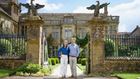 Luke and Julie Montagu standing between the stone gateposts in front of Mapperton House - a sandstone Jacobean manor