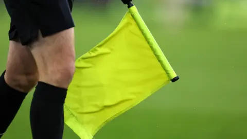 Getty Images The legs of a football referee who is wearing black socks and holding a yellow flag. The grass is blurred in the background. 