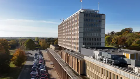 A large office block in a rectangular shape can be seen with cars parked below on a sunny day