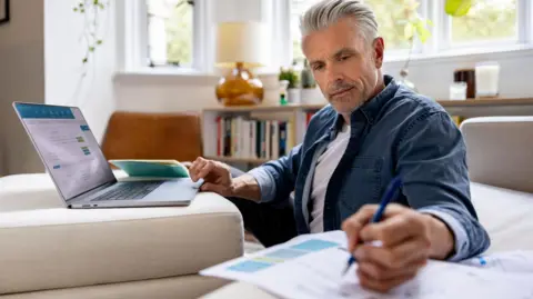 Man sitting behind a laptop with bills to one side in a living room.