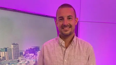 Jake Barrett is photographed at the BBC WM studio. There is a screen behind him showing a view of Birmingham and the wall is lit up in purple. Mr Barrett has short hair and a beard. He is wearing a light shirt and trousers.