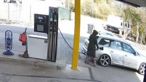 Mari Elin Jones CCTV footage showing a black-hooded thief wearing red and white pyjama trousers filling up the tank of their silver car. It's a sunny day and an empty forecourt.