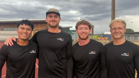 Four men standing on a running track. They are all wearing long-sleeve black tops with a white and orange Miles That Matter logo on the chest. The man on the left has black curly hair. The man next to him has brown facial hair and is wearing a camouflage cap. Next to him is a man with brown curly hair and facial hair. The man on the right has short, blond hair. The sky is cloudy and grey.