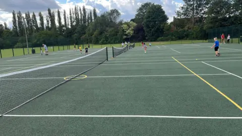 John Devine/BBC About ten youngsters aged from 6-8 playing tennis and practising shots on three hard courts with green base and white lines.