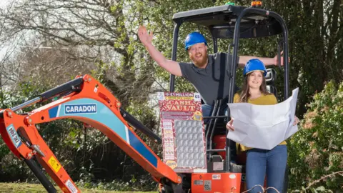 National Lottery Zoe and Craig wearing hard hats that are blue, Craig is sat in an orange digger and Zoe is holding blue prints for plans.