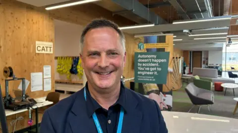 A man in a navy shirt and jacket, wearing a turquoise lanyard smiles at the camera. He has short dark grey hair and is standing in a large foyer area in front of some signage and chairs and tables.