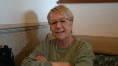 Shaun Whitmore/BBC Carmel Westgate wearing glasses and a green jumper with her arms crossed. She is looking into the camera.