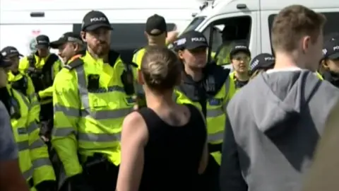 A woman in a black vest top faces police with a man in a grey hoodie stood to her right. A line of uniformed police officers stand in front of her with a van in the background.