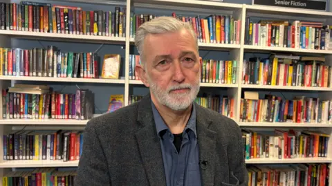 Rod McCready in a school library with books on shelves behind him. He has grey hair and a grey beard. He is wearing a grey wool suit and a blue shirt