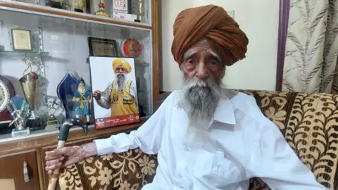 Pardeep Sharma/BBC Singh seen at his home in Beas Pind, next to a photo of him wearing a medal