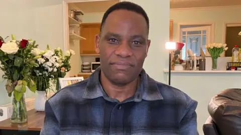 Dean wearing a dark plaid shirt sat in Shayne's family home. He is smiling at the camera. There are lots of bunches of flowers dotted around in the background. 