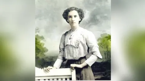 Gabrielle Deans A lady stands holding onto a chair in what appears to be a very old photograph. She is wearing a blouse and skirt and is in a photography studio with a tree lined background behind her