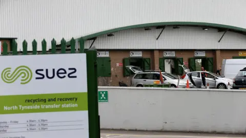 Cars line up at a recycling centre in front of green disposal hatches. There is a white, green and black recycling centre sign in the foreground.
