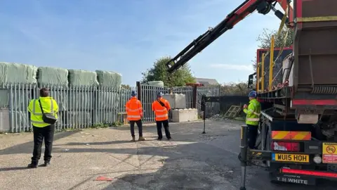 Environment Agency Four people in high-vis jacket, two orange and two yellow. They are stood on a patch of tarmac, all facing a large grey metal fence. Behind the fence are large green bales stacked on top of each other three high. To the right is a lorry. On it is a large arm which is moving two concrete blocks into place against the grey fencing.